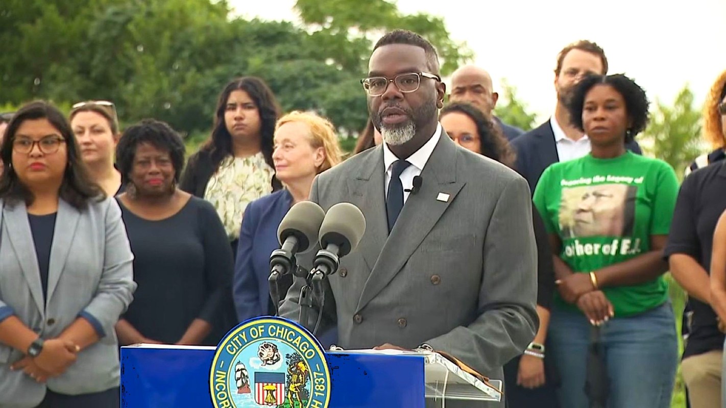 A man speaks at a podium with microphones, surrounded by a group of people.