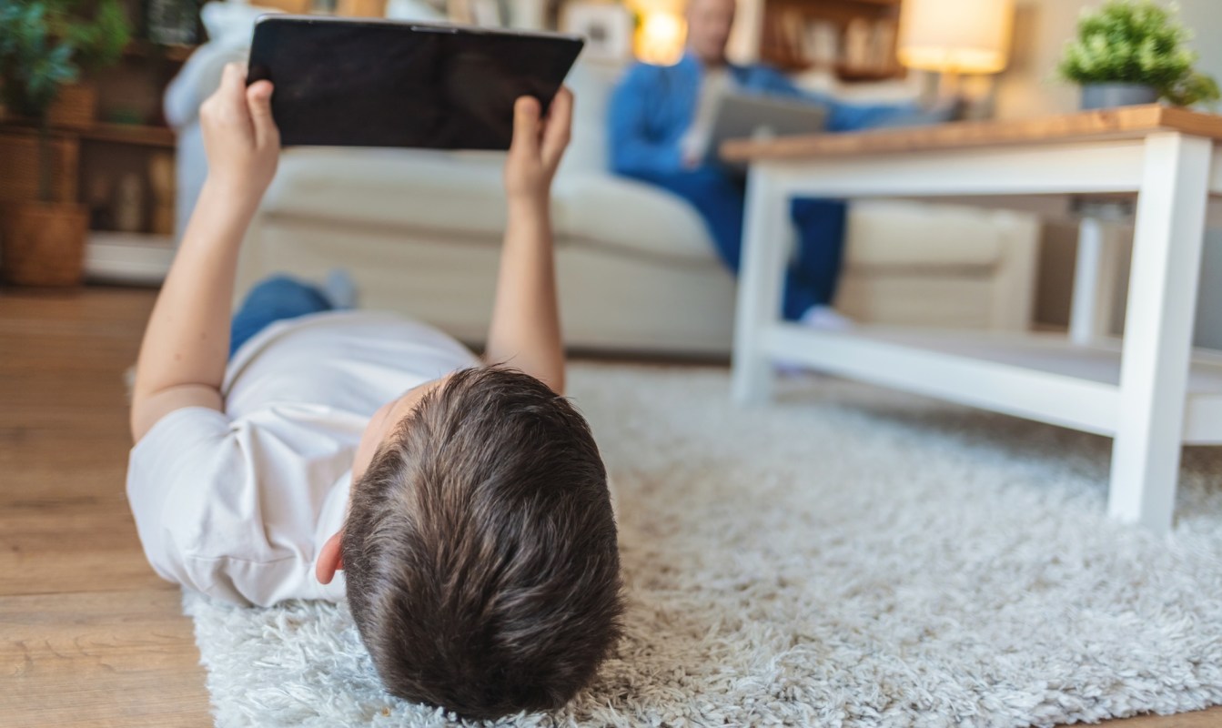 A child lying on a rug holds a tablet while an adult sits nearby on a couch.