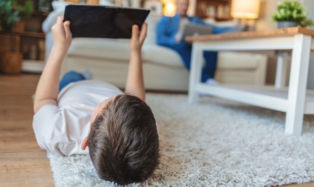 A child lying on a rug holds a tablet while an adult sits nearby on a couch.