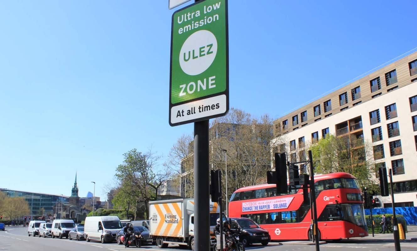 A ULEZ zone sign is displayed in London, with various vehicles and buildings in the background.