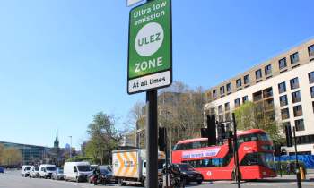 A ULEZ zone sign is displayed in London, with various vehicles and buildings in the background.