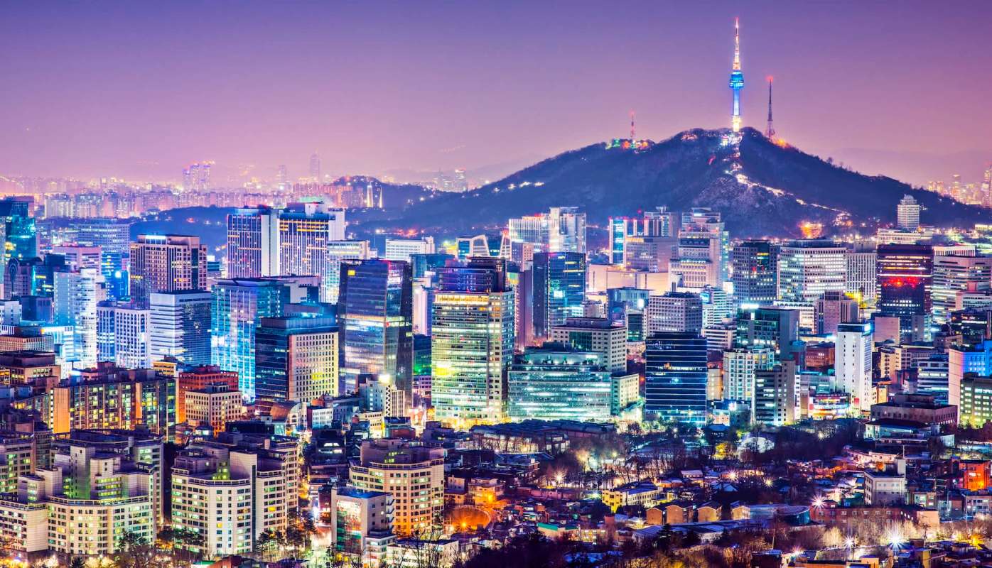 A city skyline at night with brightly lit buildings and a mountain in the background featuring a communication tower.