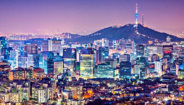 A city skyline at night with brightly lit buildings and a mountain in the background featuring a communication tower.