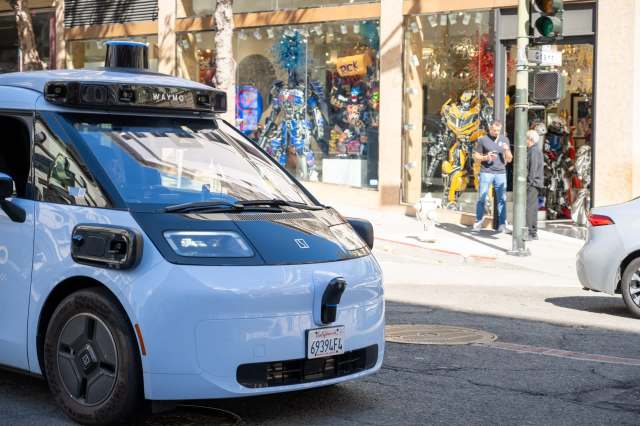 A Waymo autonomous vehicle parked on a city street with a shop displaying colorful merchandise in the background.