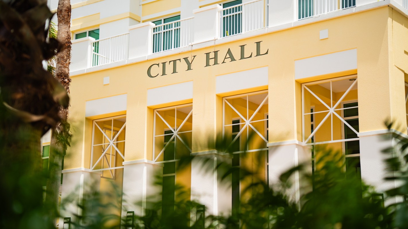 A city hall building with yellow walls and large windows, partially obscured by greenery.