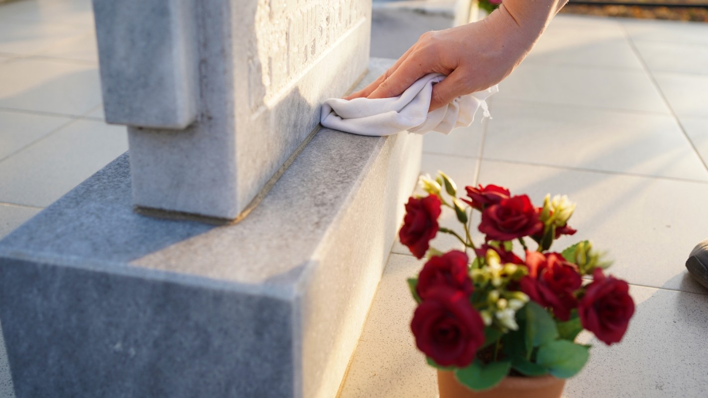 A hand wipes a gravestone near a bouquet of red roses on a tiled surface.