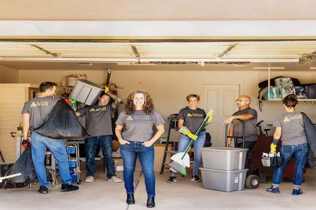 A group of six people in a garage, wearing matching shirts, with cleaning supplies and bins.