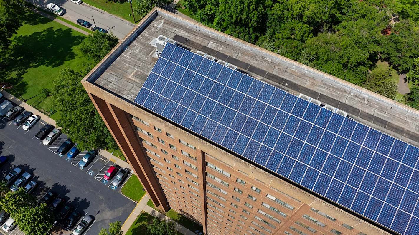 A rooftop covered in solar panels on a multi-story brick building, with a parking lot and trees below.
