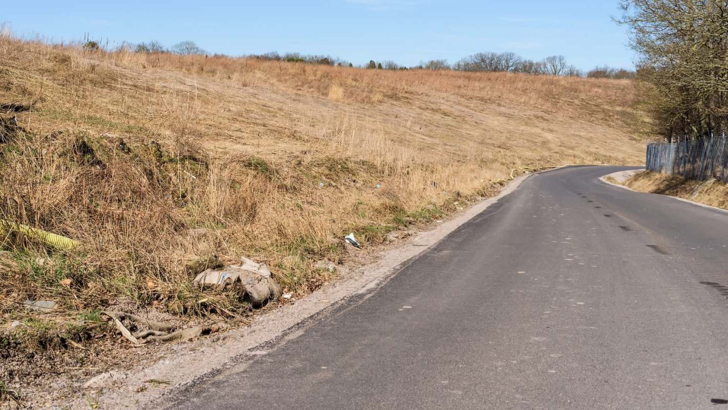 A winding road alongside the hillside of a landfill covered with dry grass and scattered debris.