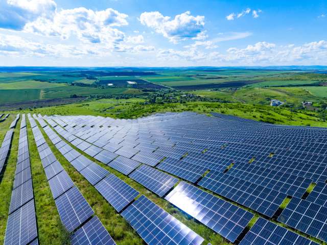 A solar panel field covering a green hillside under a bright blue sky with scattered clouds.