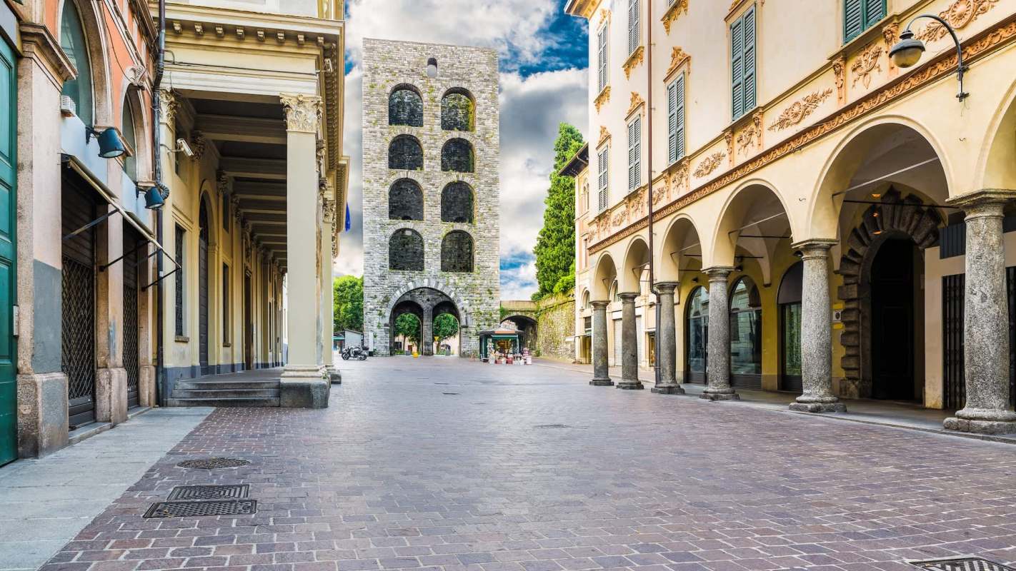 A historic stone tower stands at the end of a cobblestone street lined with arches and buildings.