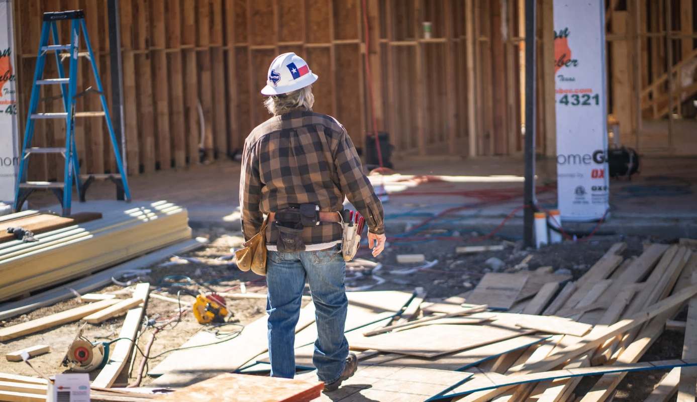 A construction worker with a Texas flag on his helmet stands on a site with wood and tools around at a construction site.