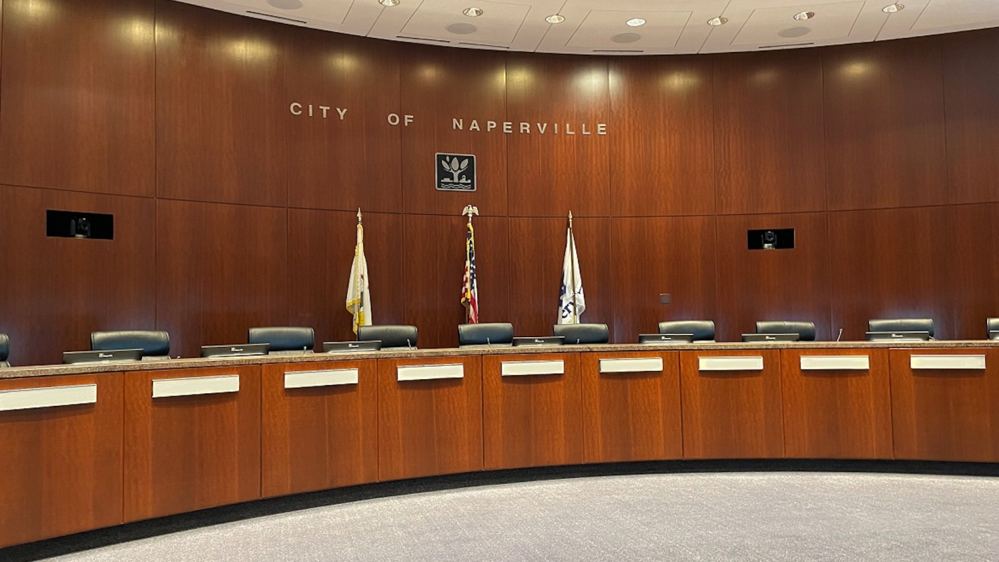 A wooden council chamber with several empty chairs, flags, and the words "CITY OF NAPERVILLE" on the wall.