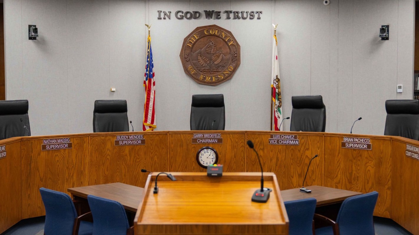 A empty government meeting room with a wooden table, chairs, and a seal on the wall.