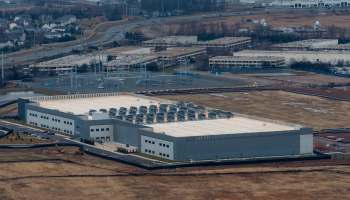 A large industrial building with numerous cooling units on the roof, surrounded by open land and roads.