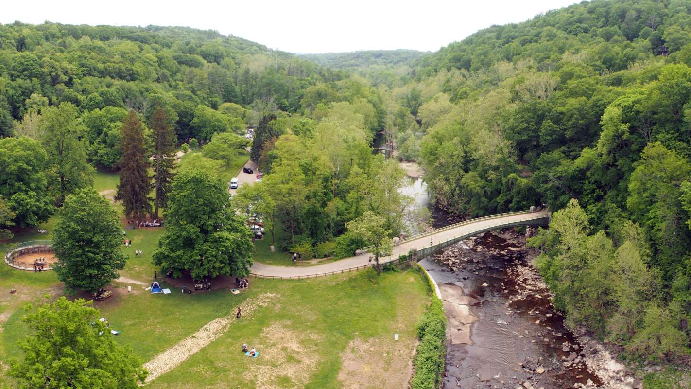 An aerial view of a park with green trees, a river, and a bridge, featuring people and cars nearby.