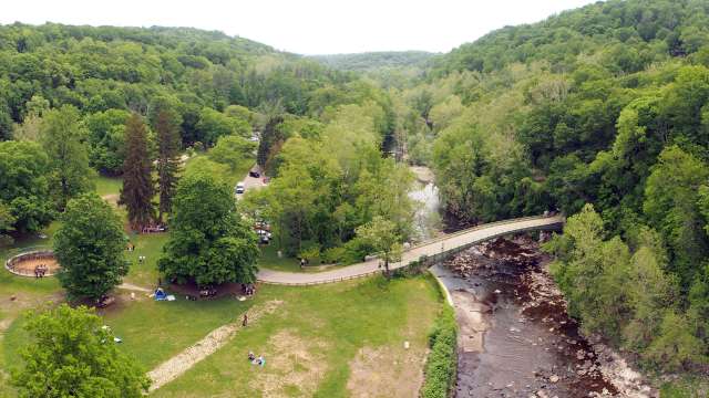 An aerial view of a park with green trees, a river, and a bridge, featuring people and cars nearby.