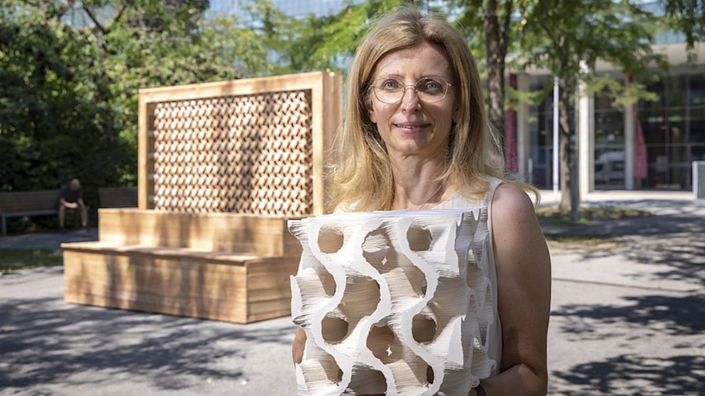 A woman holds a sculptural object in front of a geometric wooden installation in a park.