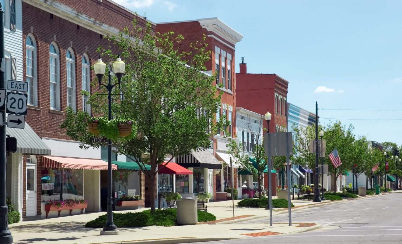 A street view of a small town featuring shops with awnings and decorative trees along the sidewalk.