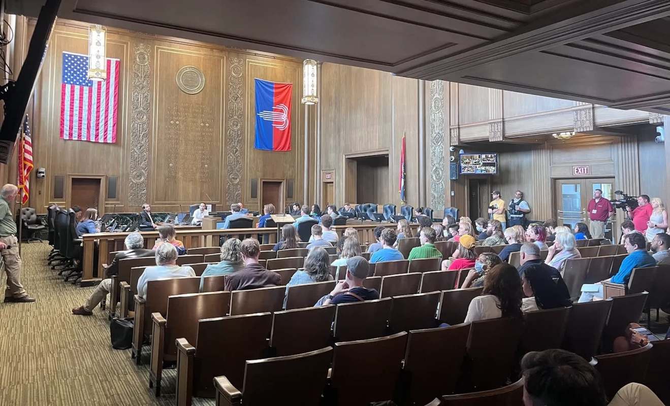 A public meeting in a large room with people seated, facing a panel of speakers and decorated with flags.