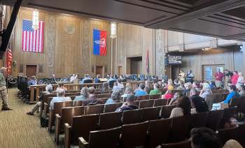 A public meeting in a large room with people seated, facing a panel of speakers and decorated with flags.