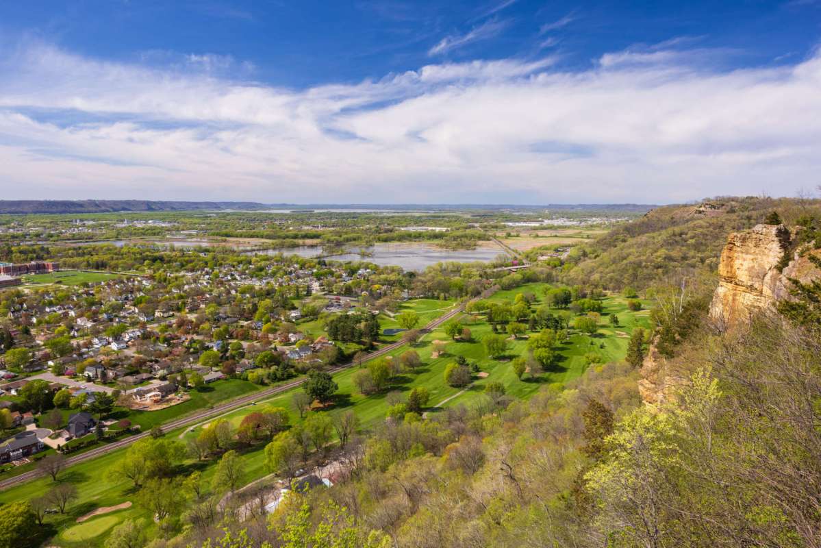 A panoramic view of a landscape featuring greenery, a river, and distant hills under a blue sky.
