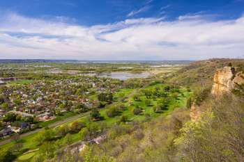 A panoramic view of a landscape featuring greenery, a river, and distant hills under a blue sky.
