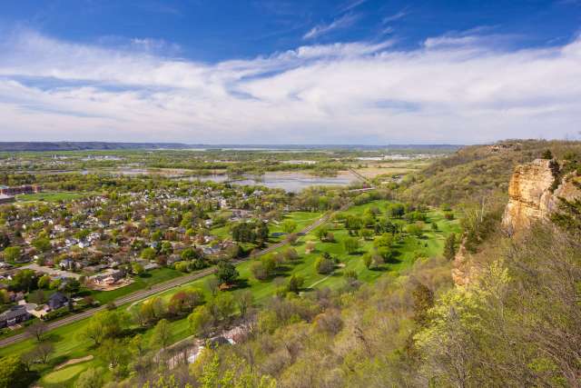 A panoramic view of a landscape featuring greenery, a river, and distant hills under a blue sky.