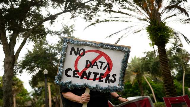A person holds a sign that reads "NO DATA CENTERS" in a park with palm trees.