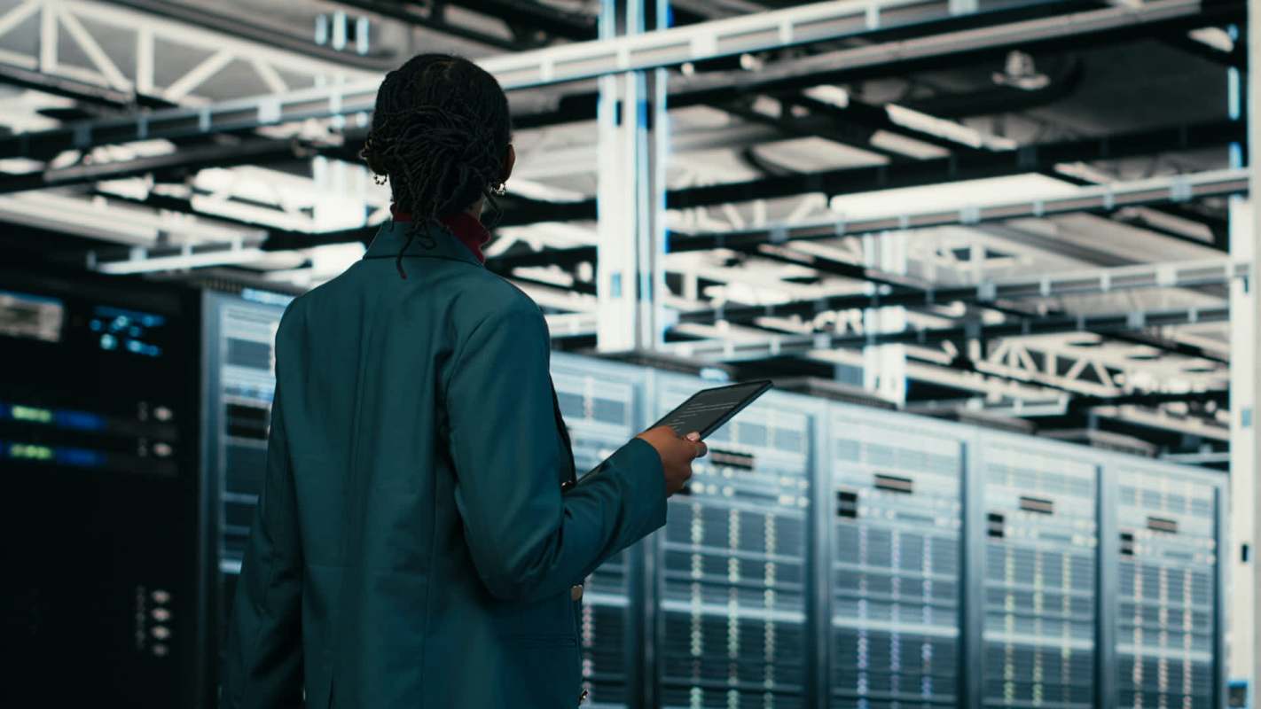 A person holds a tablet while standing in front of server racks in a data center.