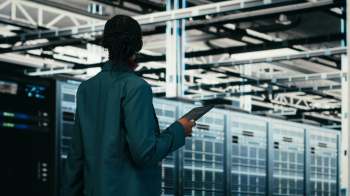 A person holds a tablet while standing in front of server racks in a data center.