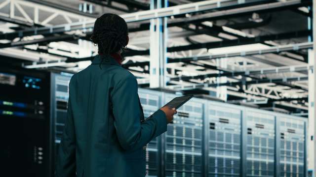 A person holds a tablet while standing in front of server racks in a data center.