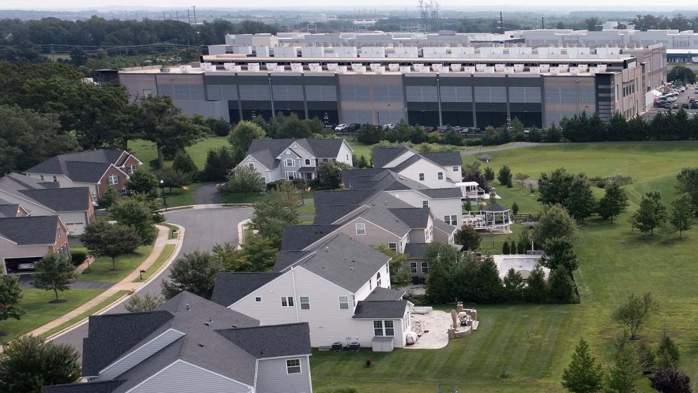 A residential neighborhood with houses, lawns, and a large industrial building in the background.