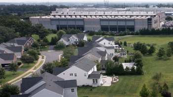 A residential neighborhood with houses, lawns, and a large industrial building in the background.