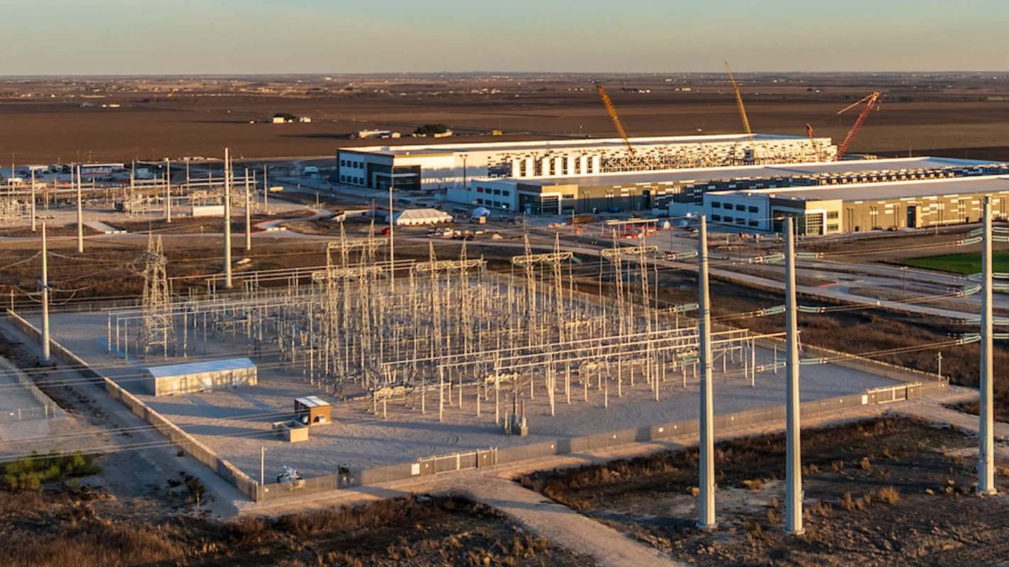 An electrical substation with power lines, surrounded by flat terrain and a large building in the background.
