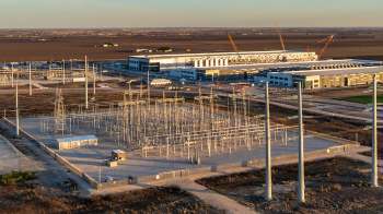 An electrical substation with power lines, surrounded by flat terrain and a large building in the background.