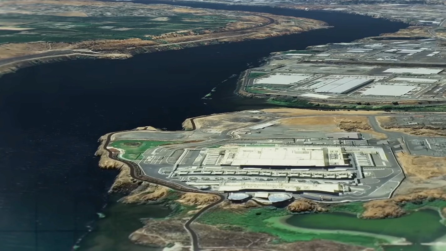 An aerial view of data center buildings next to a river, surrounded by land and vegetation.