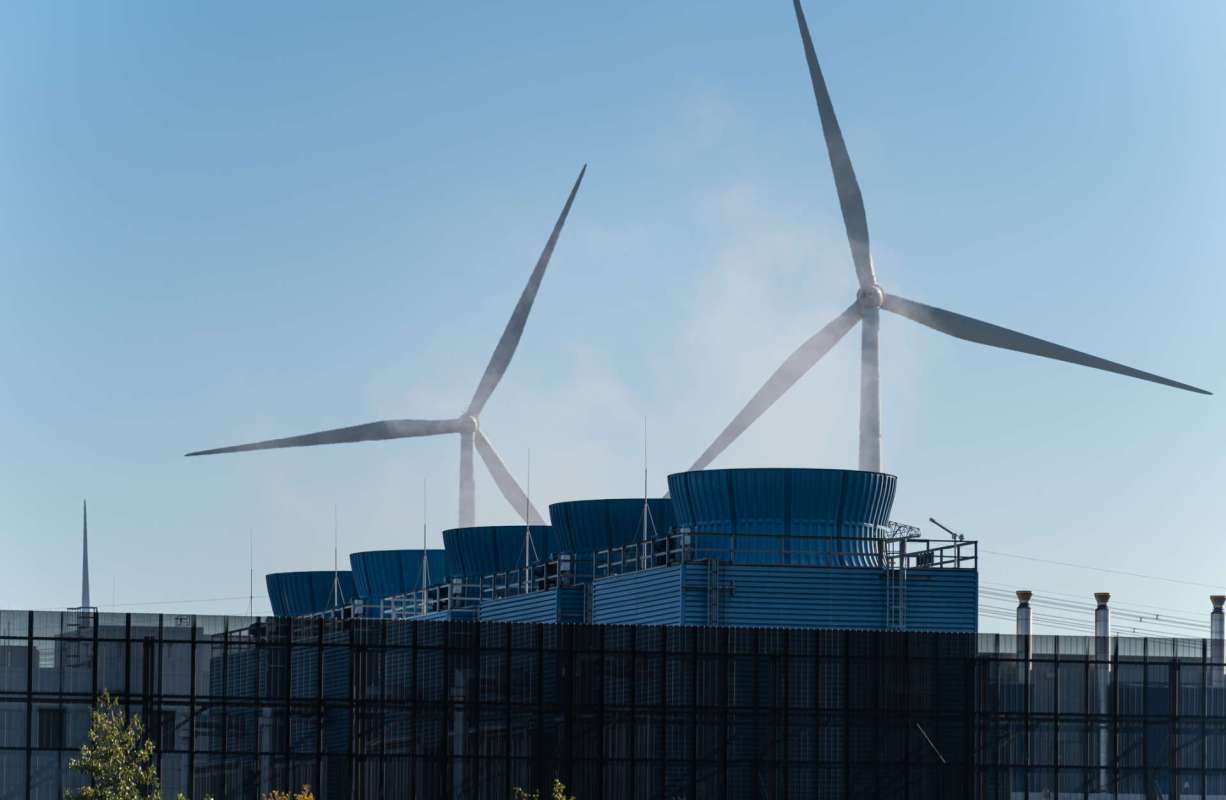 Two wind turbines behind cooling towers emitting vapor against a clear blue sky.