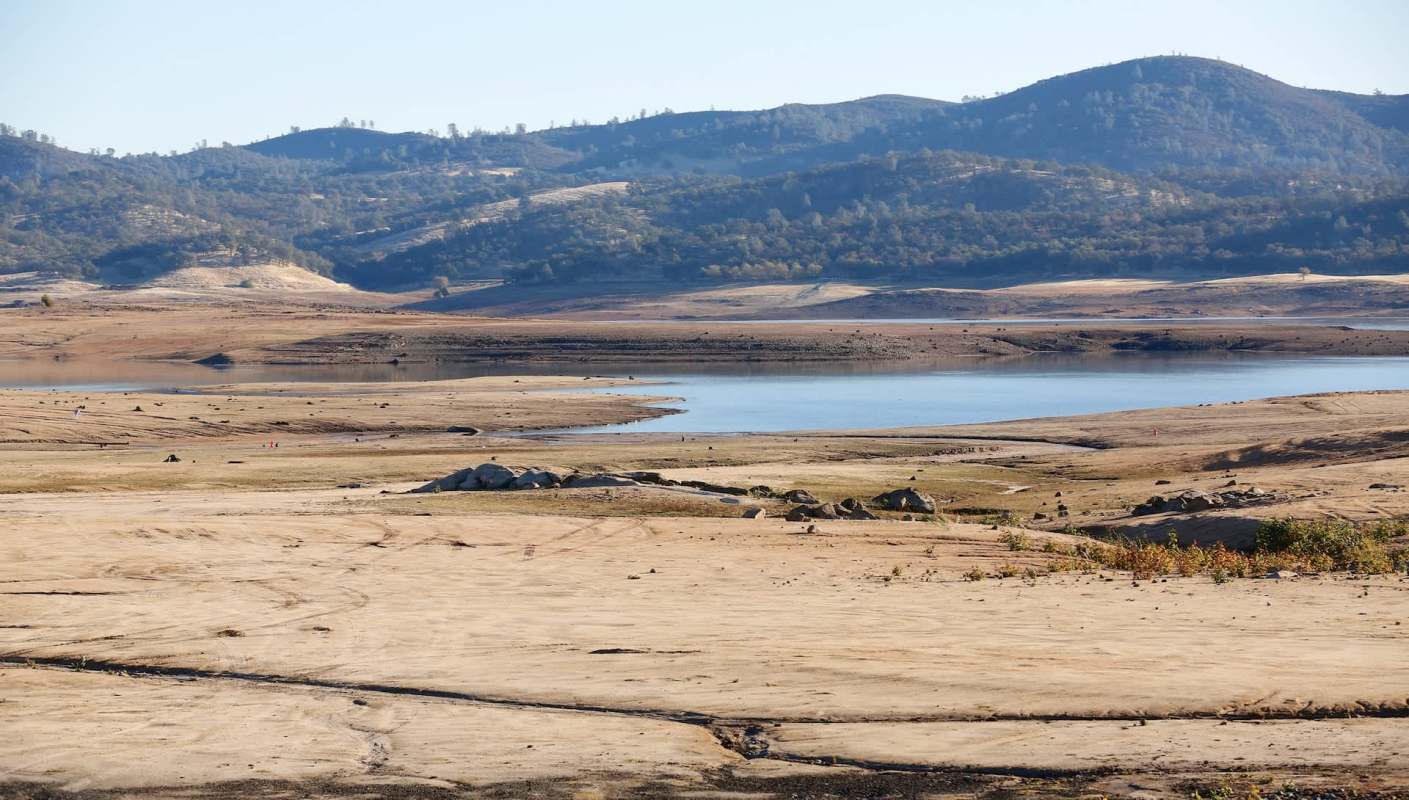 A dry landscape featuring a lake, rocky shore, and distant mountains under a clear blue sky.