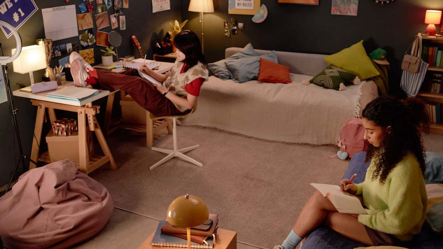 Two young women are working on creative projects in a cozy room with a sofa, desk, and decorative items on the walls.