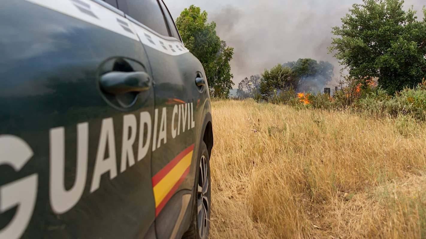 A civil guard vehicle is parked near a field with visible smoke and flames from a fire.