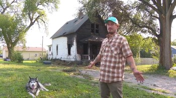 A man stands in front of a damaged house with a husky lying on the grass nearby.