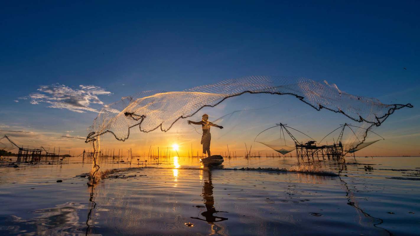 A person casting a fishing net into calm water during sunset, with fishing equipment visible in the background.