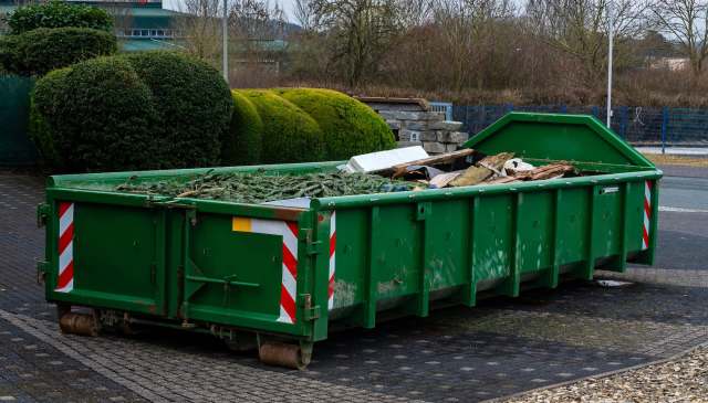 A green dumpster filled with debris, including branches and wood, situated on a paved surface.