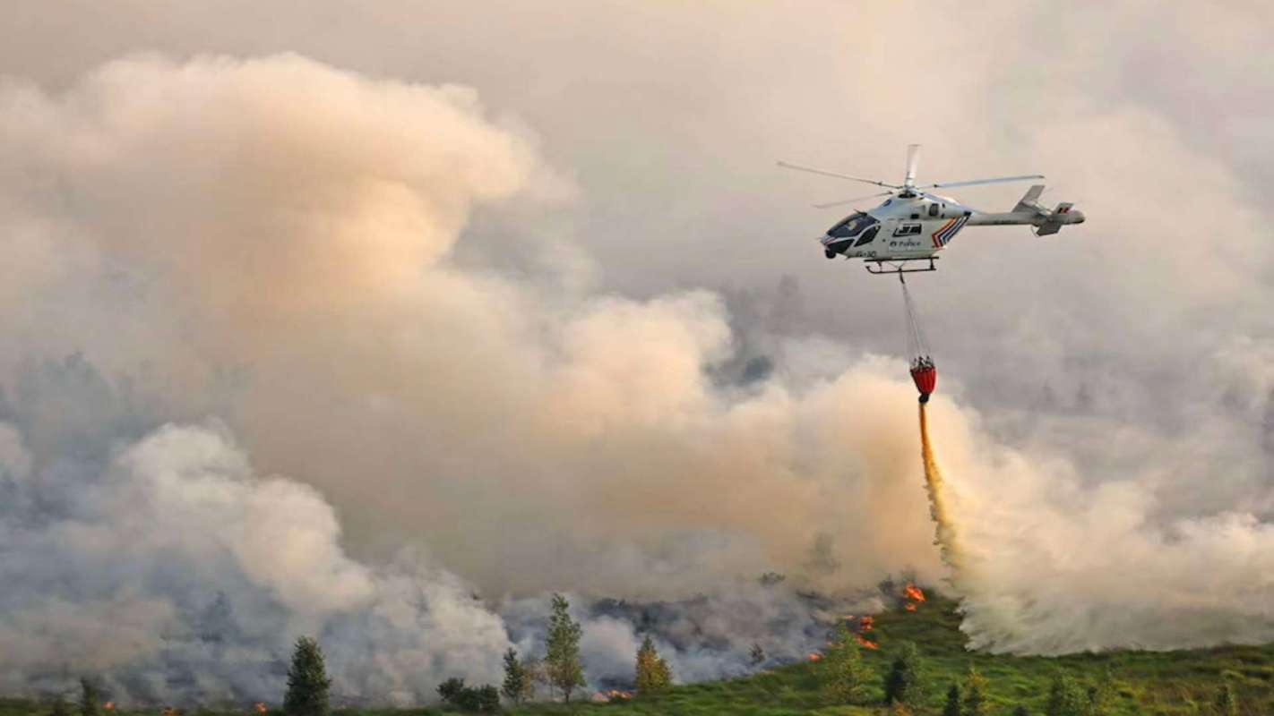 A helicopter drops water on a forest fire, surrounded by thick smoke and flames.