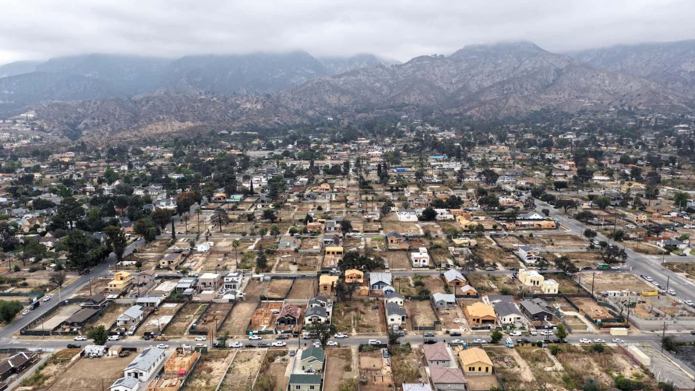 An aerial view of a residential area with houses and streets, set against a mountainous backdrop.