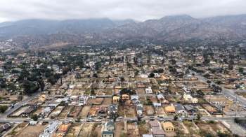 An aerial view of a residential area with houses and streets, set against a mountainous backdrop.