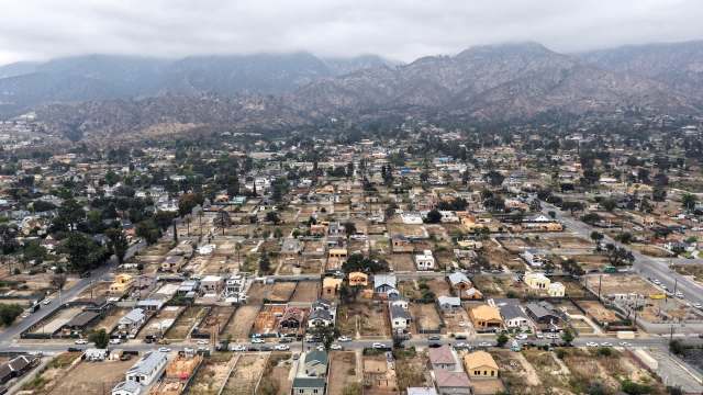 An aerial view of a residential area with houses and streets, set against a mountainous backdrop.