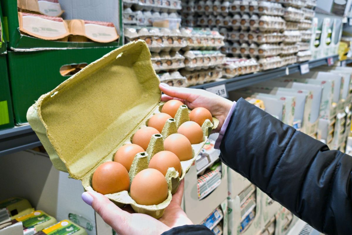 A person holds an open carton containing ten eggs in a grocery store aisle.