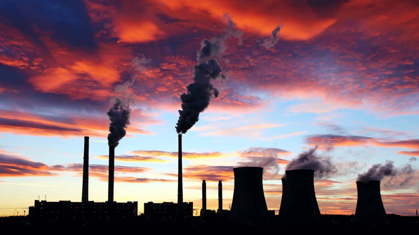 Industrial smokestacks emit smoke against a colorful sunset sky.